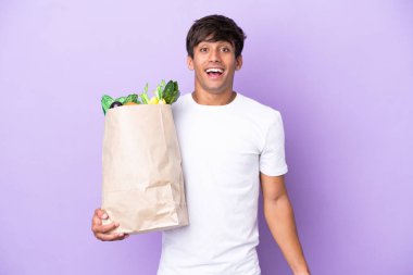 Elinde bir alışveriş çantası tutan genç adam mor arka planda, şaşırmış bir yüz ifadesiyle izole edilmiş.