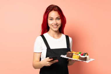 Pasta şefi pembe arka planda izole edilmiş bir kek tutuyor ve cep telefonuyla bir mesaj gönderiyor.