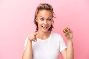 Elinde Bitcoin tutan genç bir Rus kız pembe arka planda izole edilmiş şaşırmış ve ön tarafı işaret ediyor.