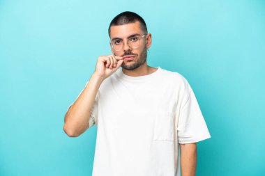 Mavi arka planda izole edilmiş genç beyaz bir adam sessizlik işareti gösteriyor.