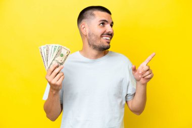 Kafkasyalı genç adam sarı arka planda izole edilmiş bir sürü para alıyor. Harika bir fikir.