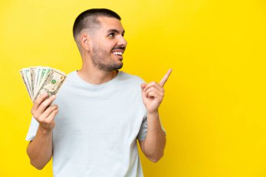 Genç beyaz adam sarı arka planda izole edilmiş bir sürü para alıyor. Çözümü bulmak için parmağını kaldırıyor.