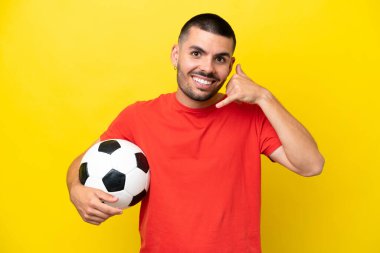 Futbol oynayan beyaz bir genç sarı arka planda izole edilmiş bir şekilde telefon jesti yapıyor. Bana geri dönüş işareti ver.