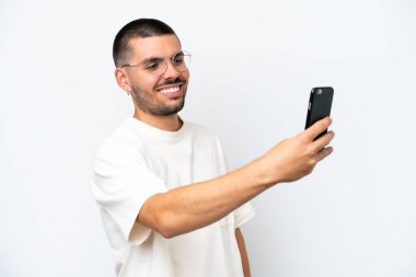 Beyaz arka planda izole edilmiş genç beyaz bir adam cep telefonuyla selfie çekiyor.