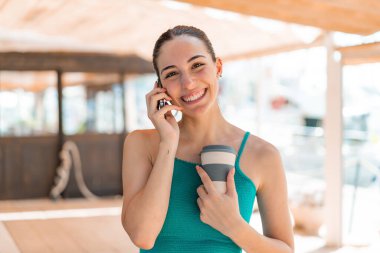 Açık havada cep telefonu kullanan ve elinde mutlu bir ifadeyle kahve tutan genç ve güzel bir kadın.