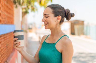 Genç ve güzel bir kadın açık havada elinde kahveyle mutlu bir ifadeyle.