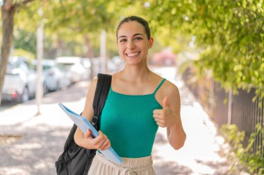 Açık havada genç ve güzel bir kız öğrenci. Başparmakları havada. Çünkü iyi bir şey oldu.