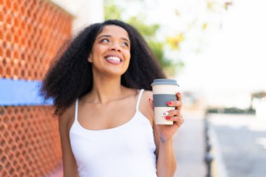 Genç Afrikalı Amerikalı bir kadın açık havada kahve alıp gülümseyerek yukarı bakıyor.