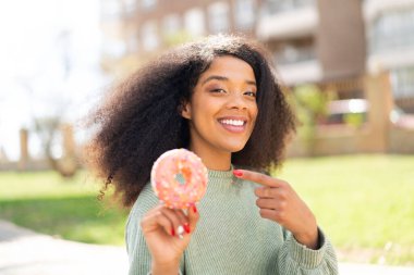 Genç Afrikalı Amerikalı bir kadın açık havada donut tutuyor ve ona doğrultuyor.