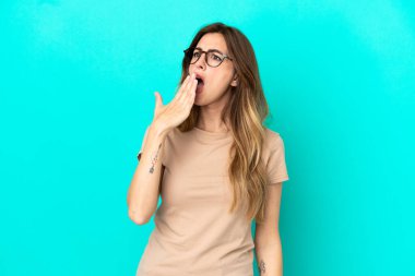 Genç beyaz kadın mavi arka planda izole edilmiş esniyor ve geniş bir ağzı eliyle kapatıyor.