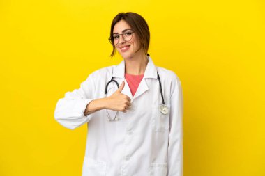 Doktor kadın sarı arka planda izole edilmiş, baş parmak işareti yapıyor.