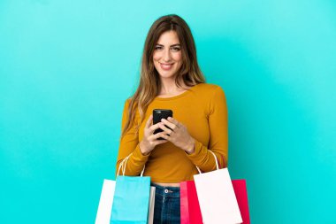 Beyaz bir kadın mavi arka planda izole edilmiş. Elinde alışveriş poşetleri var ve bir arkadaşına cep telefonuyla mesaj yazıyor.