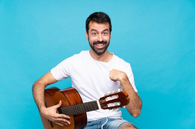 İzole edilmiş mavi arka planda gitar çalan genç bir adam. Sürpriz bir yüz ifadesi var.