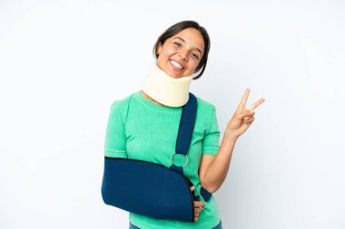 Beyaz arka planda izole edilmiş boyunluk takan genç İspanyol kadın gülümsüyor ve zafer işareti gösteriyor.