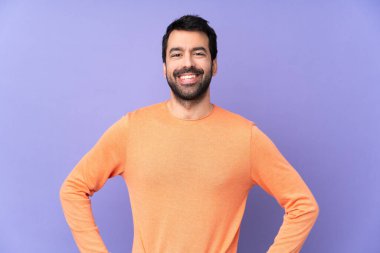 Mor arka planda, kolları kalçasında poz veren ve gülümseyen beyaz tenli yakışıklı bir adam.