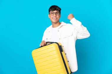 Young man over isolated blue background in vacation with travel suitcase