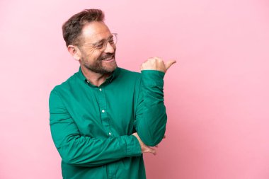 Pembe arka planda izole edilmiş orta yaşlı beyaz bir adam bir ürün sunmak için kenarı işaret ediyor.