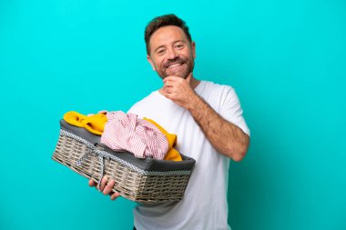 Mavi arka planda soyutlanmış bir çamaşır sepetini tutan orta yaşlı kadın mutlu ve gülümsüyor.