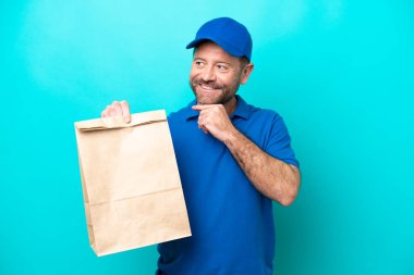 Middle age man taking a bag of takeaway food isolated on blue background looking to the side and smiling