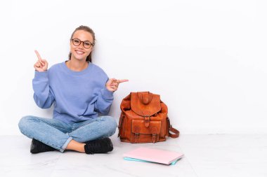 Beyaz arka planda izole bir katta oturan genç kız parmak uçlarını işaret ediyor ve mutlu.