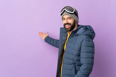 İzole edilmiş mor arka planda snowboard gözlüklü kayakçı. Gelmesi için ellerini yana uzatıyor.