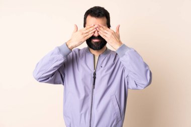 Sakallı, ceketli, soyutlanmış arka planda beyaz bir adam. Gözleri elleriyle kaplıyor ve gülümsüyor.