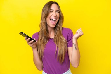 Sarı arka planda izole edilmiş genç beyaz kadın cep telefonu kullanıyor ve zafer jesti yapıyor.