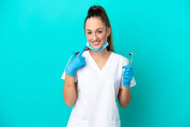 Elinde mavi arka planda izole edilmiş aletler tutan beyaz dişçi kadın baş parmak hareketi yapıyor.