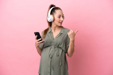 Pembe arka planda izole edilmiş genç bir kadın hamile ve işaret ederken müzik dinliyor.
