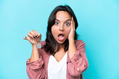 Genç İspanyol kadının elinde ev anahtarları var. Mavi arka planda, şaşırmış ve şok olmuş bir yüz ifadesi var.