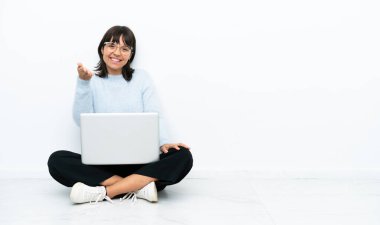 Genç melez kadın dizüstü bilgisayarla yerde oturuyor. Beyaz arka planda izole edilmiş, iyi bir anlaşma için el sıkışıyor.