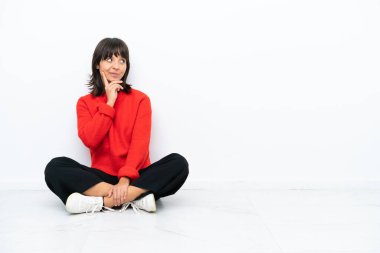 Genç melez kadın, beyaz arka planda izole bir şekilde yerde oturuyor. Şüpheleri var ve kafası karışmış bir yüz ifadesi var.