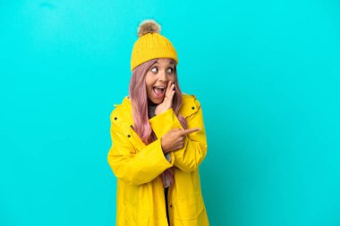 Pembe saçlı, yağmura dayanıklı bir ceket giyen, mavi arka planda izole edilmiş genç bir kadın bir ürünü sunmak ve bir şeyler fısıldamak için kenarı işaret ediyor.