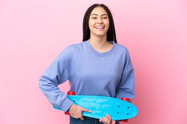 Saçı örgülü genç bir kız pembe arka planda mutlu bir ifadeyle kaykay yapıyor.