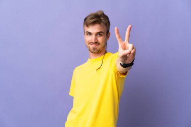 Mor arka planda izole edilmiş yakışıklı sarışın adam gülümsüyor ve zafer işareti gösteriyor.