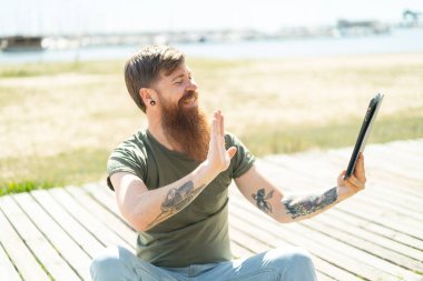 Kızıl saçlı, sakallı bir adam elinde tablet, selam veriyor.