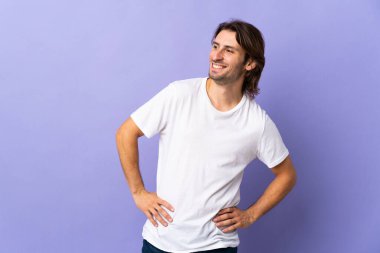 Mor arka planda soyutlanmış, kolları kalçasında ve gülümseyerek poz veren genç yakışıklı bir adam.