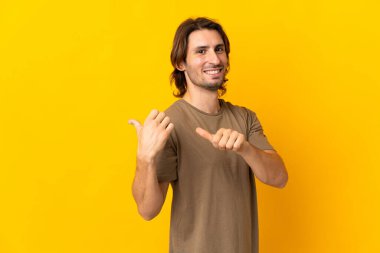 Sarı arka planda izole edilmiş genç yakışıklı bir adam bir ürün sunmak için kenarı işaret ediyor.