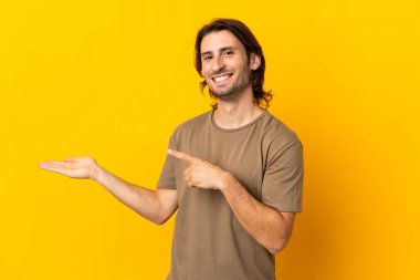 Sarı arka planda izole edilmiş genç yakışıklı bir adam. Avucunun içinde hayali bir ilan tutuyor.