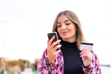 Genç, güzel Romen kadın açık havada cep telefonuyla kredi kartıyla alışveriş yapıyor.