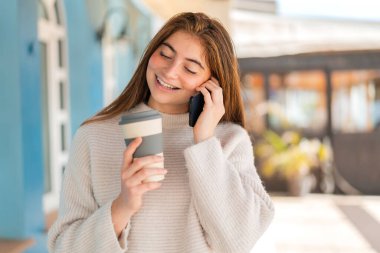 Genç beyaz bir kadın cep telefonu kullanıyor ve elinde mutlu bir ifadeyle kahve tutuyor.