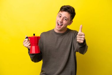 Elinde kahve demliği tutan genç Brezilyalı adam sarı arka planda izole edilmiş, başparmakları havada çünkü iyi bir şey oldu.