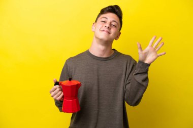 Sarı arka planda kahve demliği tutan genç Brezilyalı adam parmaklarıyla beşi sayıyor.