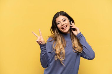 Sarı arka planda izole edilmiş cep telefonu kullanan genç Rus kız gülümsüyor ve zafer işareti gösteriyor.