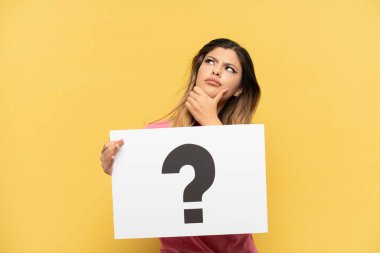 Sarı arka planda izole edilmiş genç bir Rus kızı soru işareti olan bir pankart tutuyor ve düşünüyor.