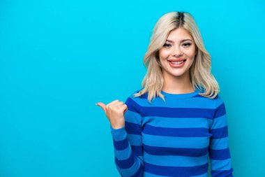 Mavi arka planda izole edilmiş genç bir Rus kadın bir ürünü sunmak için kenarı işaret ediyor.