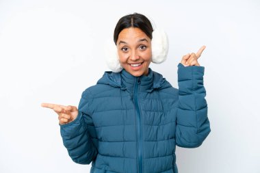 Kış kulaklığı takan genç İspanyol kadın beyaz arka planda izole edilmiş, parmak uçlarını işaret ediyor ve mutlu.