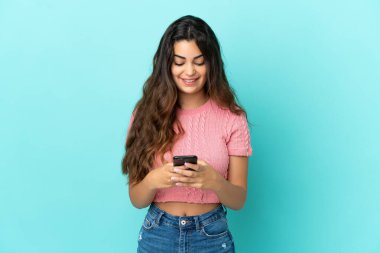 Mavi arka planda izole edilmiş genç beyaz kadın cep telefonuyla bir mesaj gönderiyor.