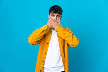 İzole edilmiş mavi arka planı olan genç bir adam elleriyle ağzını kapatıyor.
