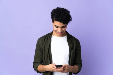 Genç Venezuelalı adam mor arka planda izole edilmiş cep telefonuyla bir mesaj gönderiyor.